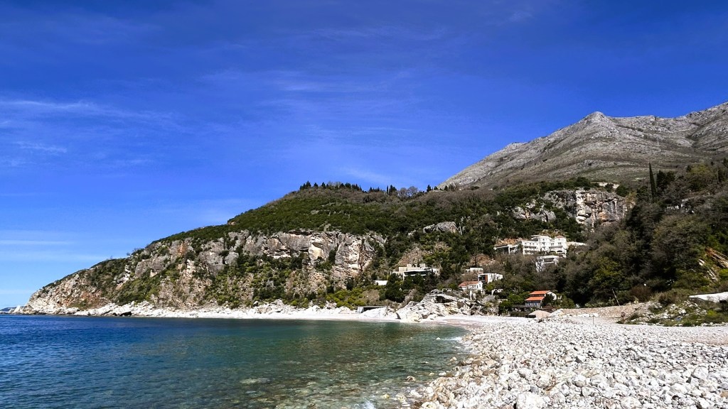 Scenic view of a rocky beach with clear blue water, surrounded by a lush green hillside and distant mountains under a bright blue sky.