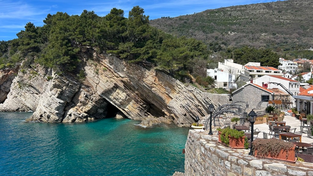 Scenic view of rocky coastline with lush trees, clear turquoise water, and a quaint seaside village featuring traditional houses and outdoor seating.