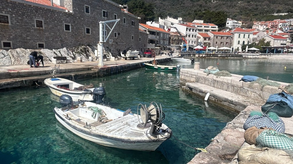 A scenic harbour with clear turquoise water, featuring several small boats docked at the pier. Stone buildings with orange roofs line the waterfront, while individuals work nearby. Green hills rise in the background, creating a picturesque coastal scene.