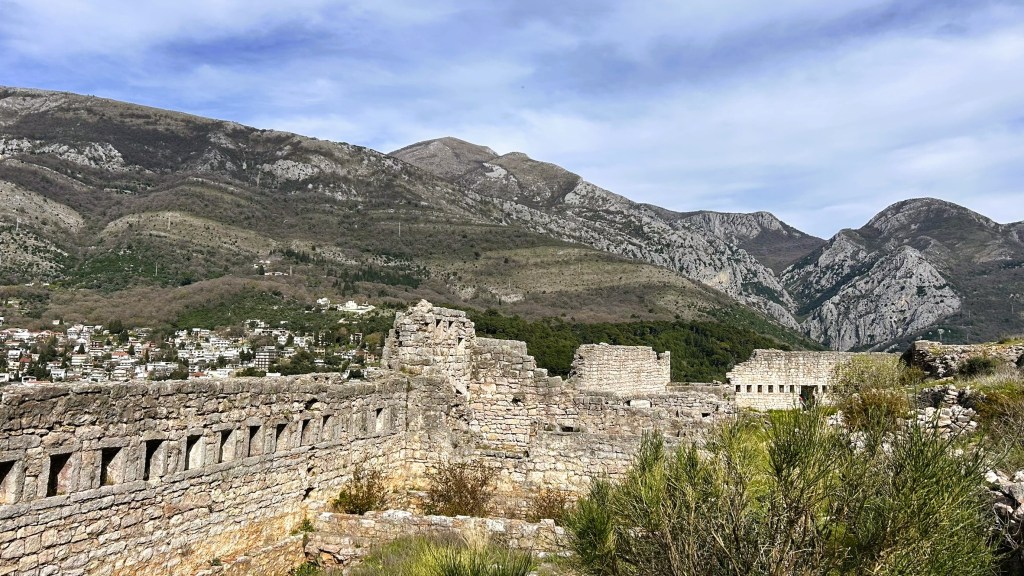 A panoramic view of ancient stone ruins set against a backdrop of rolling mountains and a small town visible in the valley.