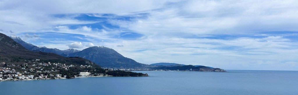Panoramic view of a coastline with mountains in the background, featuring calm blue waters and scattered clouds in the sky.