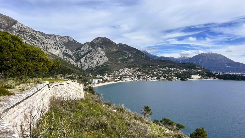A panoramic view of a coastal town surrounded by mountains, with clear blue waters in the foreground and a partly cloudy sky.