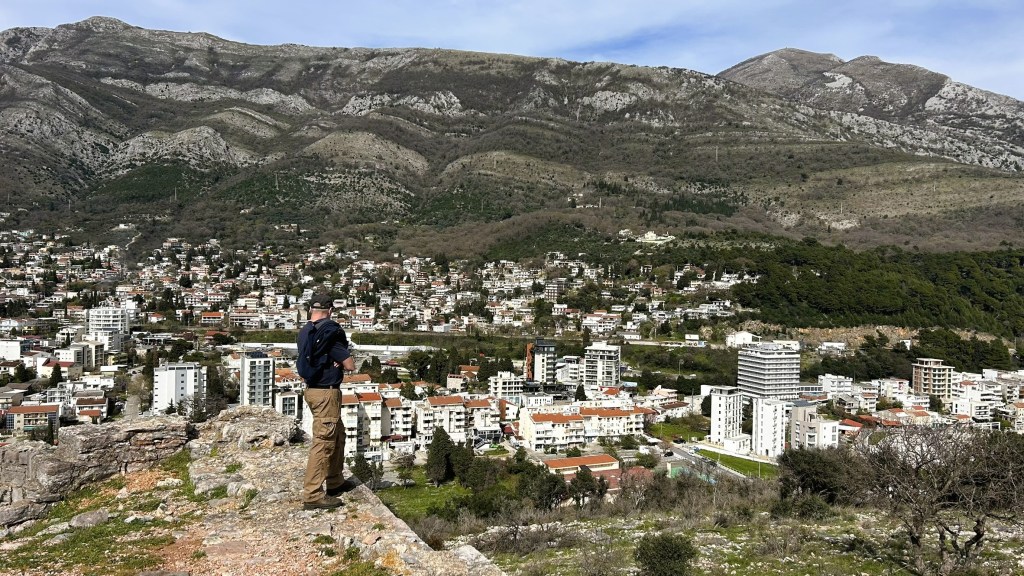 A person stands on a rocky ledge overlooking a town with modern buildings and terracotta-roofed houses, set against a backdrop of mountains under a partly cloudy sky.