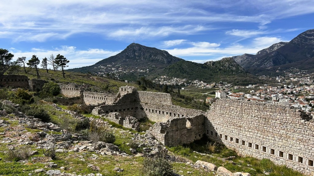 Ruins of an ancient stone structure set against a backdrop of mountains and a residential area under a clear blue sky.