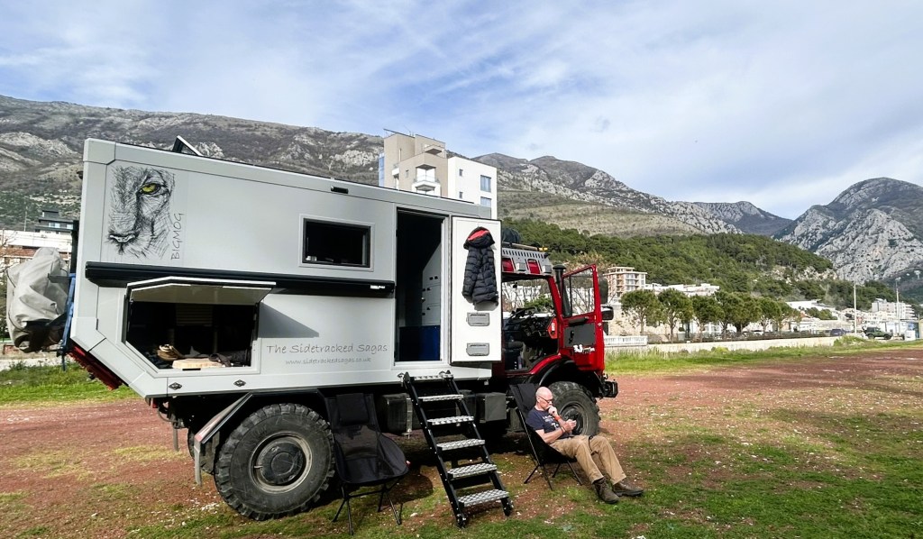 A man sitting outside a custom camper vehicle in a scenic mountainous area, with green trees and buildings in the background.