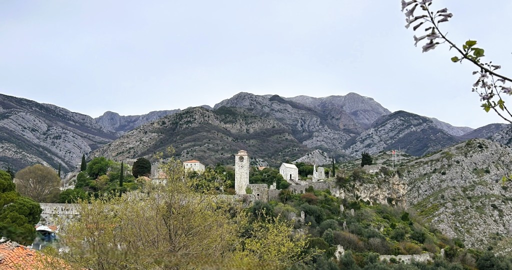 Scenic view of a mountainous landscape featuring ruins and a clock tower, with trees and village structures in the foreground.