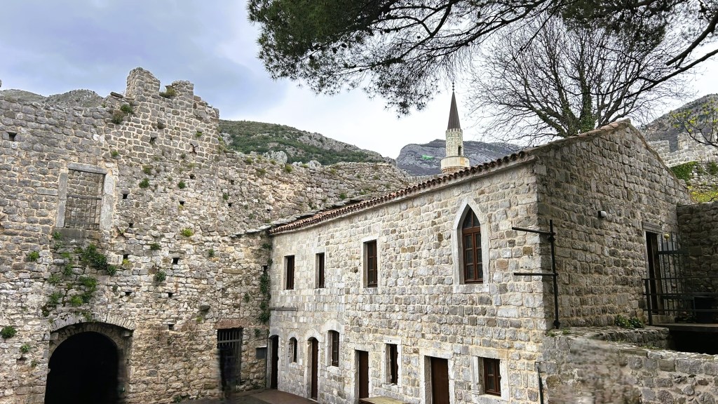Historic stone buildings and ruins with a tall, narrow spire in the background, set against a mountainous landscape under a cloudy sky.