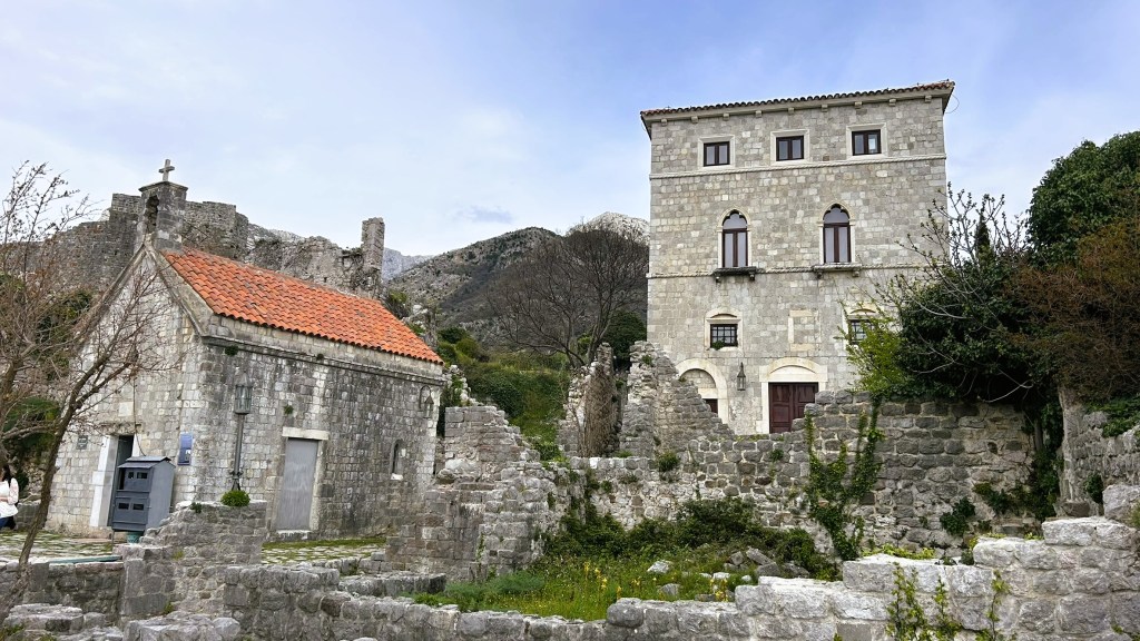 Historical stone structures and ruins in a scenic landscape, featuring a building with a red-tiled roof and a nearby tower against a mountainous backdrop.