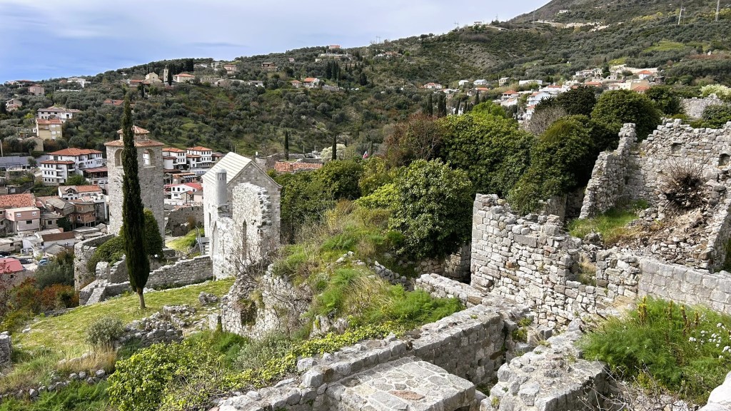 A scenic view of ancient stone ruins on a hillside, surrounded by greenery and residential buildings in the background.