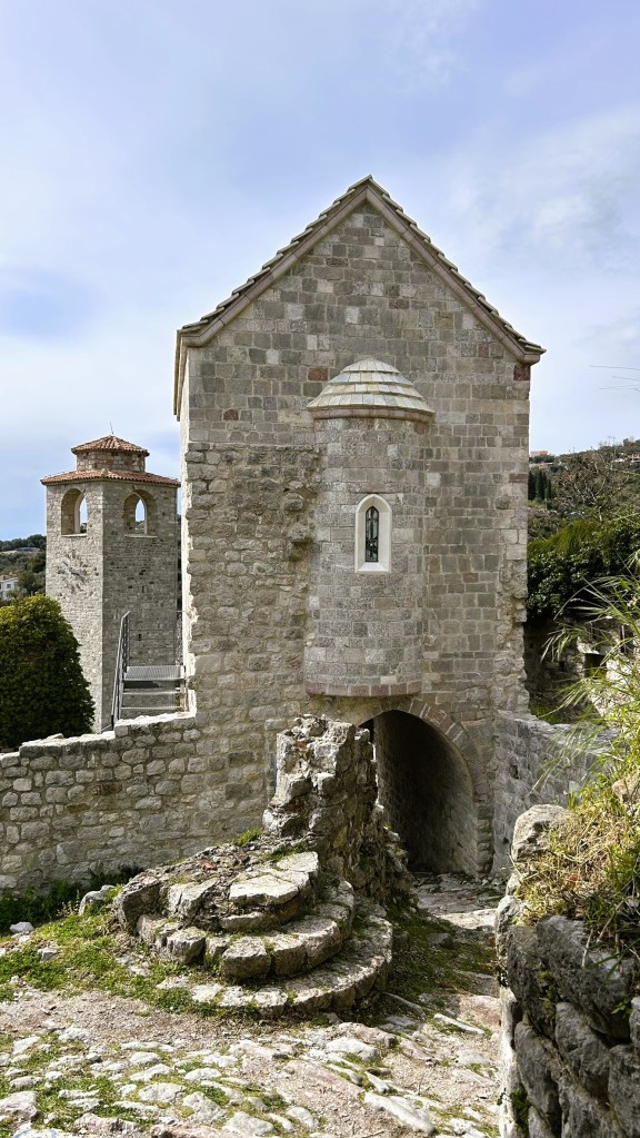 Historic stone building with a distinctive rooftop, adjacent to a bell tower, surrounded by a rocky pathway and greenery.
