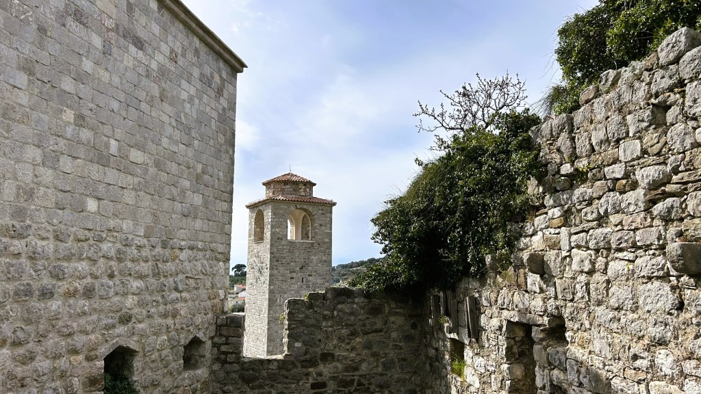 A view of ancient stone walls with a bell tower in the background, surrounded by greenery and blue skies.