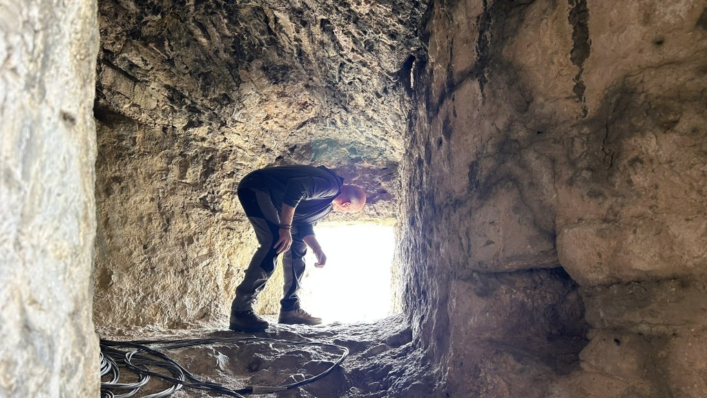 A person bent over inside a cave-like structure, examining the ground, with bright light visible at the exit.