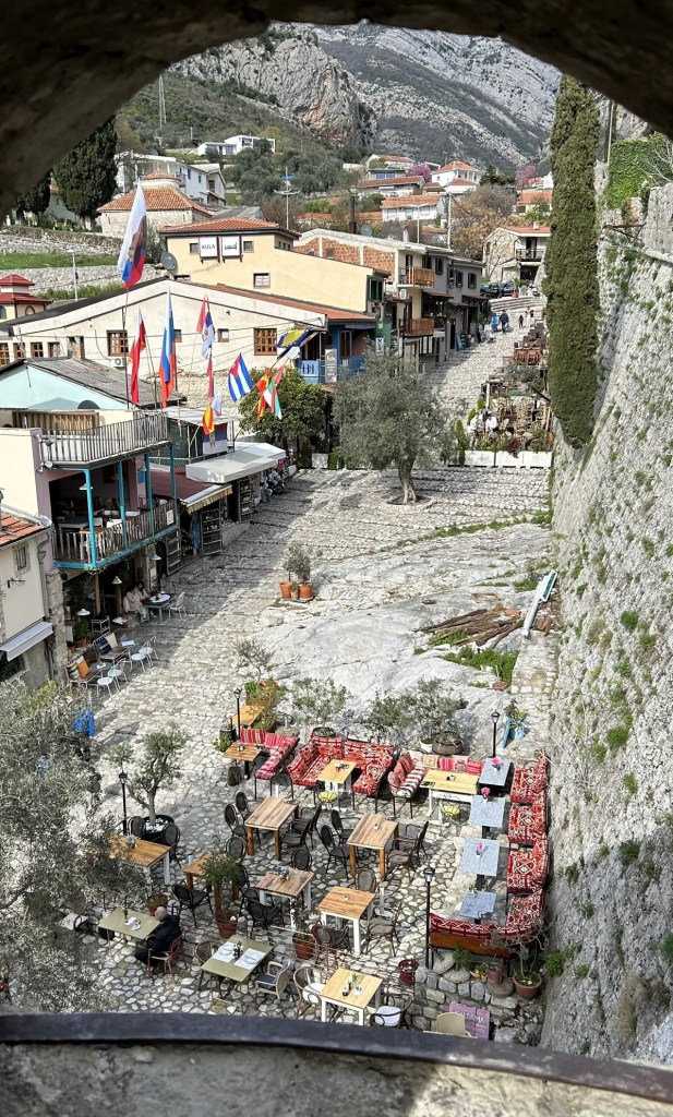 A view of a cobblestone street lined with outdoor dining areas at a quaint restaurant, featuring tables and chairs surrounded by potted olive trees. Traditional flags hang above, and buildings with rustic architecture are visible in the background.