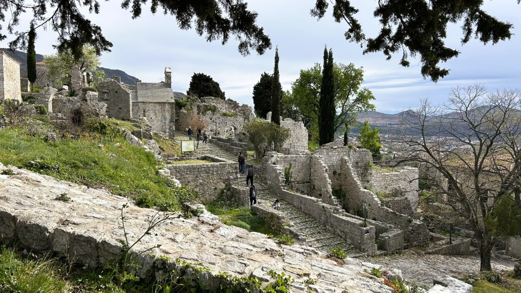 Ruins of an ancient stone structure surrounded by greenery, with people walking along stone pathways.
