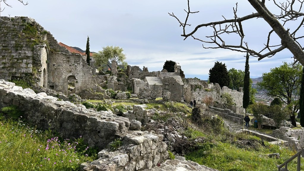 View of ancient ruins with stone structures and remains, surrounded by greenery and trees under a cloudy sky.
