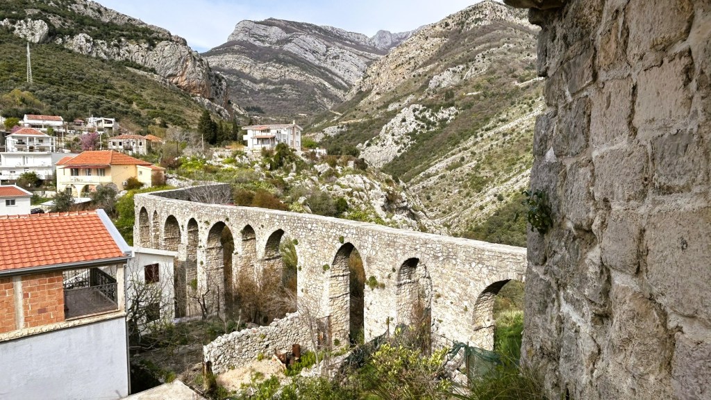A scenic view of a stone aqueduct with multiple arches, surrounded by green hills and mountains, and residential buildings in the foreground.