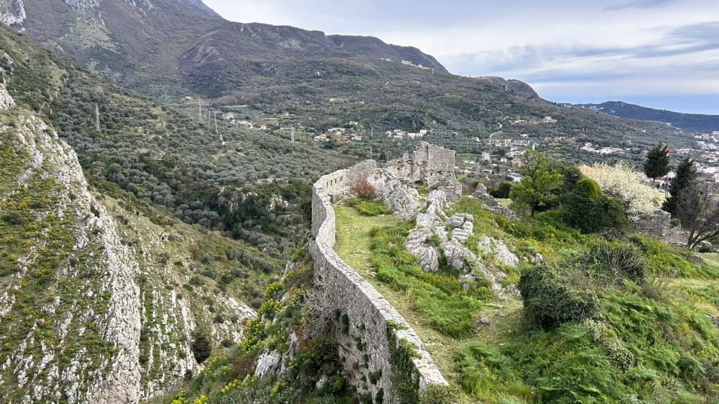 Ruins of a stone wall on a hillside surrounded by greenery and mountains under a cloudy sky.