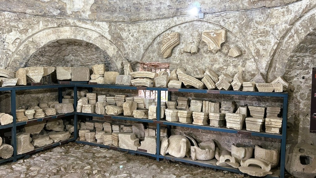 An interior view of a stone room displaying various archaeological fragments on metal shelves, including columns, capitals, and decorative elements against a rough stone wall.