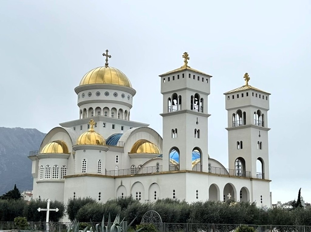 A grand church with golden domes and bell towers, set against a cloudy sky, surrounded by green foliage.