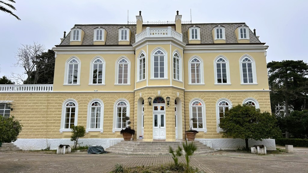 A grand yellow mansion with multiple large windows, featuring a symmetrical façade and a decorative roof, set in a landscaped area.