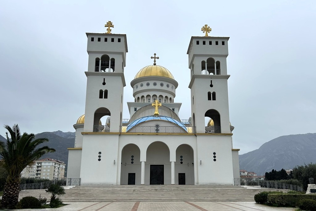 A large, modern church with golden domes and two bell towers, set against a cloudy sky and mountains in the background.