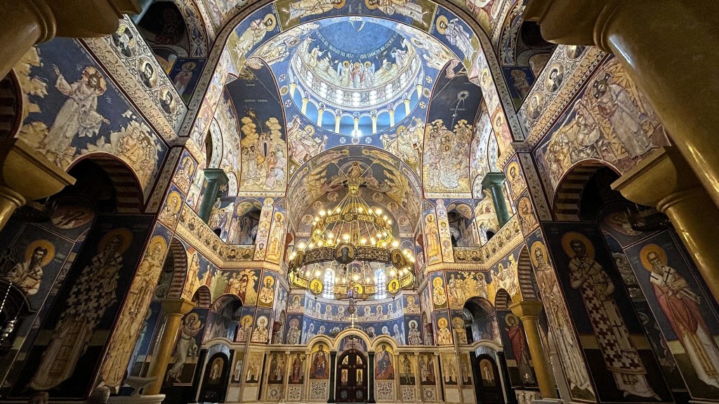 Interior view of a beautifully decorated church featuring ornate frescoes on walls and ceiling, with a large chandelier hanging in the central area.