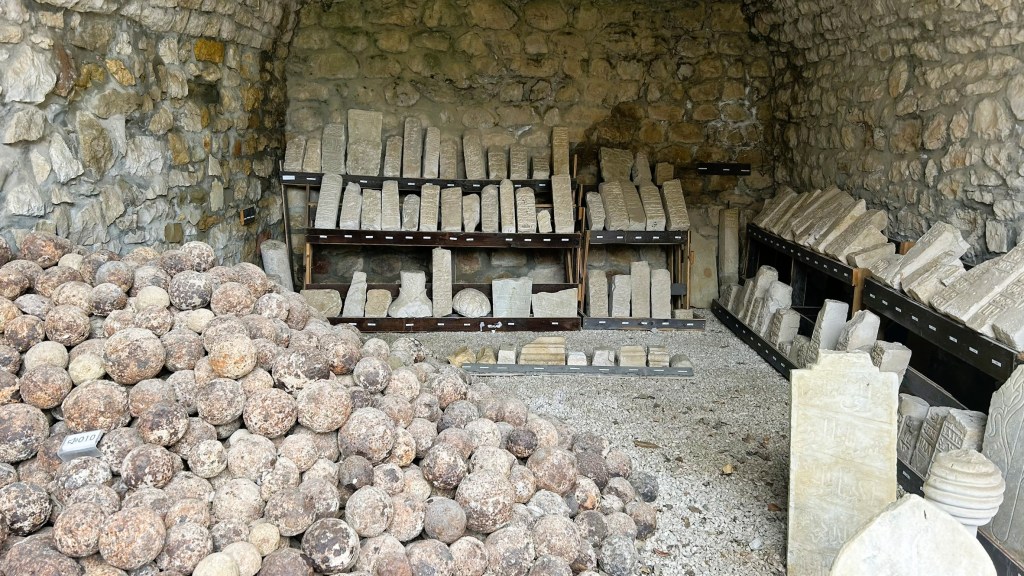 An interior view of a stone storage room featuring a pile of round rocks in the foreground and shelves filled with various stone slabs and artefacts along the walls.