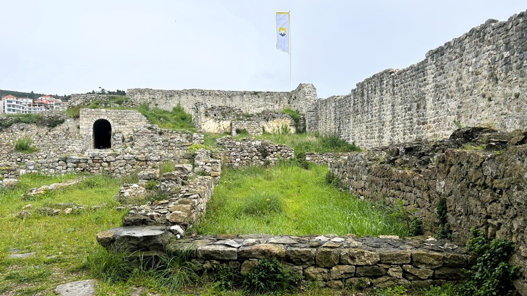 A view of ancient stone ruins with a grassy area, featuring a flag in the background and remnants of walls and archways.