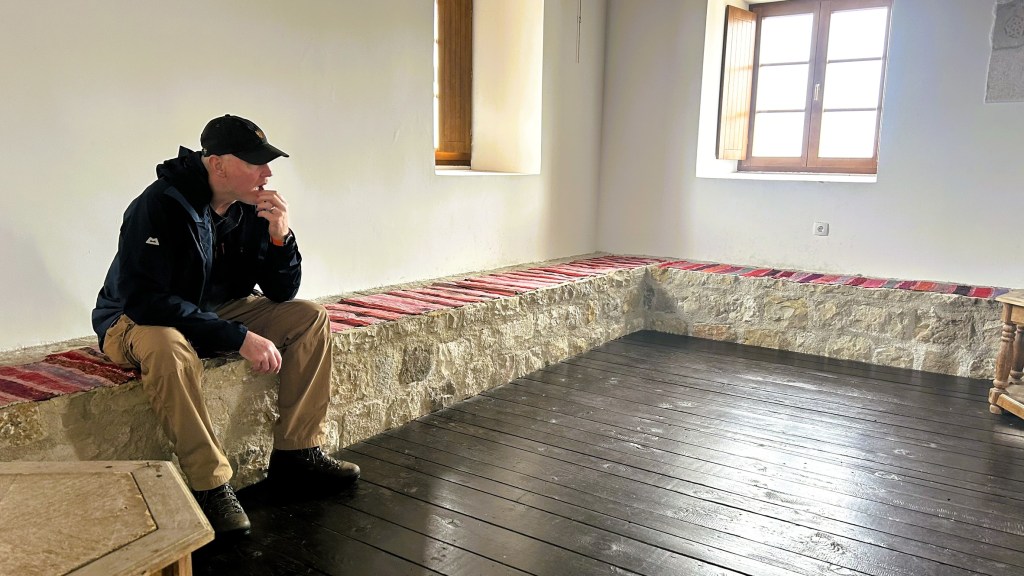 A man sitting thoughtfully on a stone bench, dressed in outdoor clothing, in a bright, minimalist room with wooden flooring and large windows.
