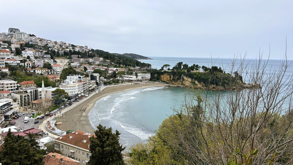 A panoramic view of a coastal town with beach, sea and hilly landscape, featuring buildings, trees and cloudy skies.