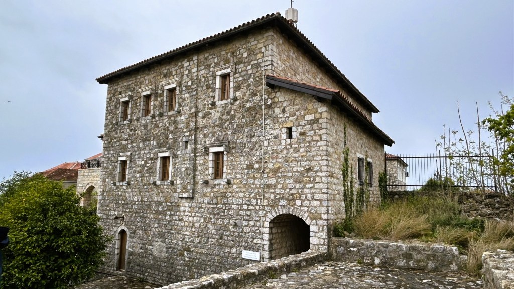 A stone building with multiple windows and a sloped roof, surrounded by greenery and a stone pathway.