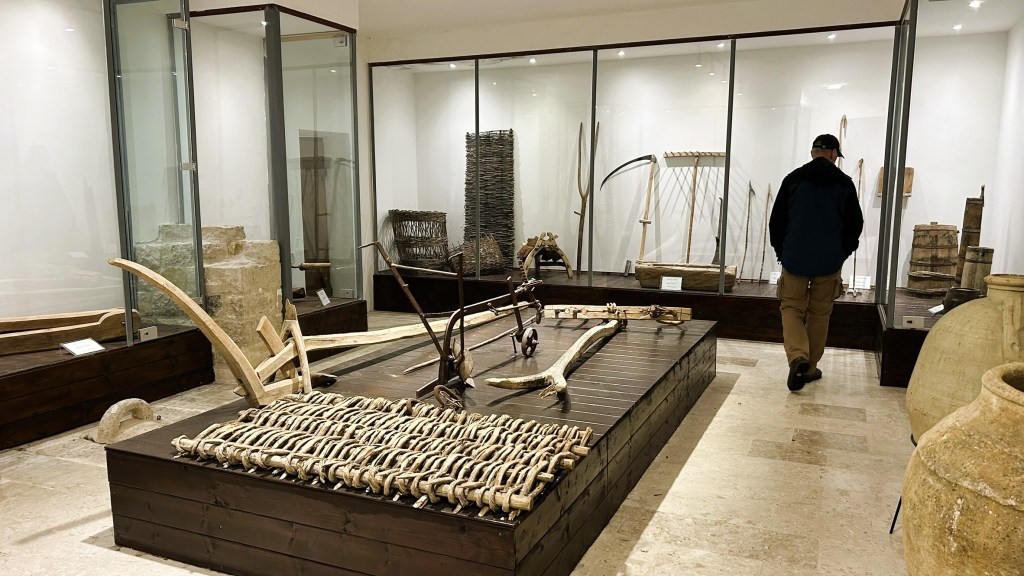 A person walking through an exhibition space showcasing various historical agricultural and crafting tools, displayed in glass cases and on wooden platforms.