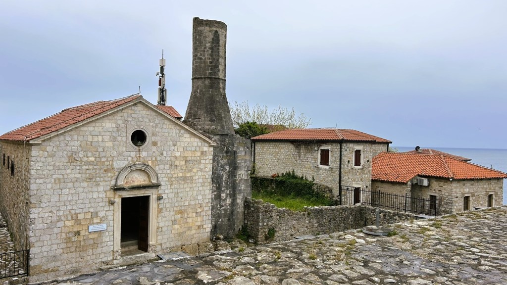 A view of a stone building with a red-tiled roof and a tall chimney, surrounded by other stone structures and greenery, set against a cloudy sky.