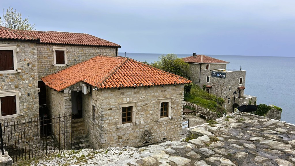 Historic stone buildings with red tiled roofs overlooking the sea, with a sign indicating a hotel nearby.