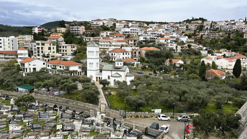 A panoramic view of a hillside town featuring residential buildings with red roofs, a church in the foreground surrounded by olive trees, and a cemetery in the lower part of the image.