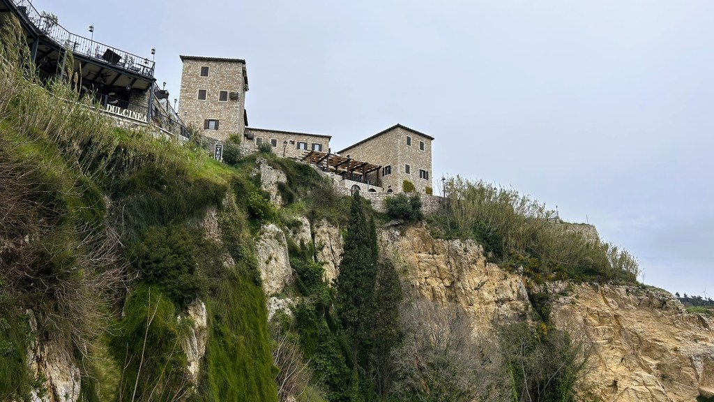 Historic stone buildings perched on a rocky cliff, surrounded by greenery and overlooking the landscape, under a cloudy sky.