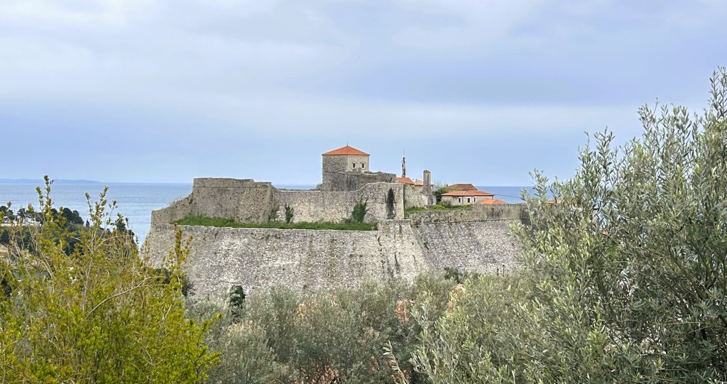 A historic stone fortress overlooking the sea, surrounded by greenery and olive trees.