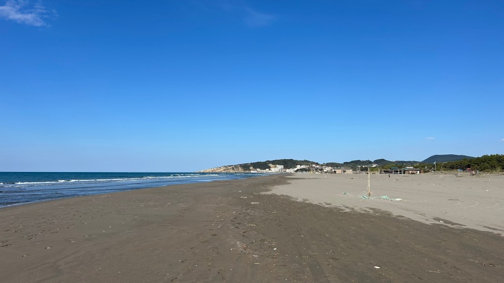 A wide sandy beach under a clear blue sky, with gentle waves lapping at the shore and a distant view of hills and buildings in the background.