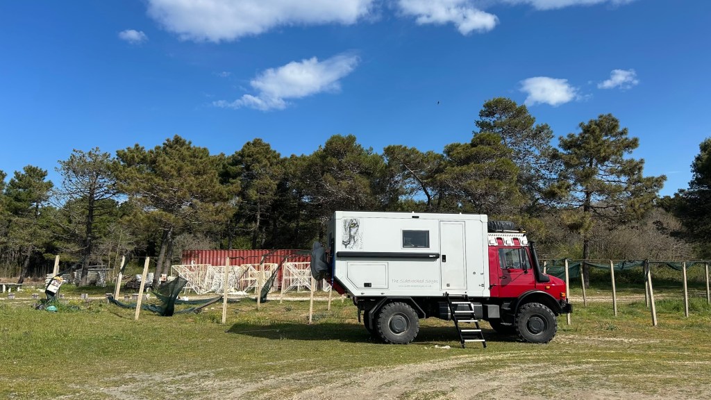 A red off-road vehicle with a white camper shell parked in a forested area, surrounded by trees and a blue sky with clouds.