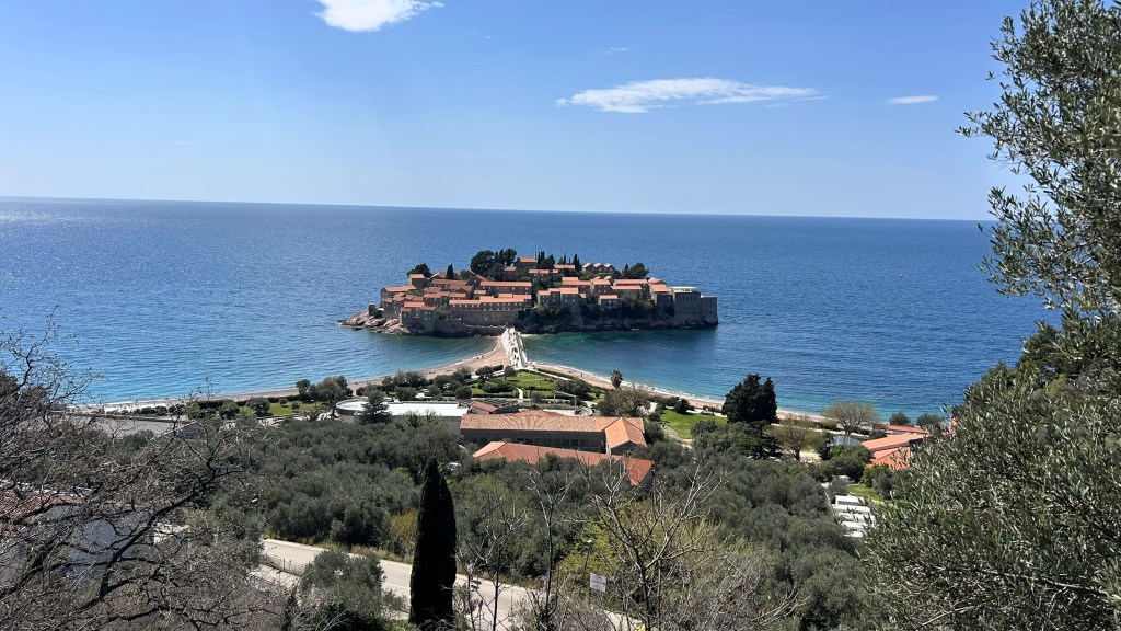 A panoramic view of a coastal island with cluster of buildings surrounded by clear blue water and a beach, under a bright sky.
