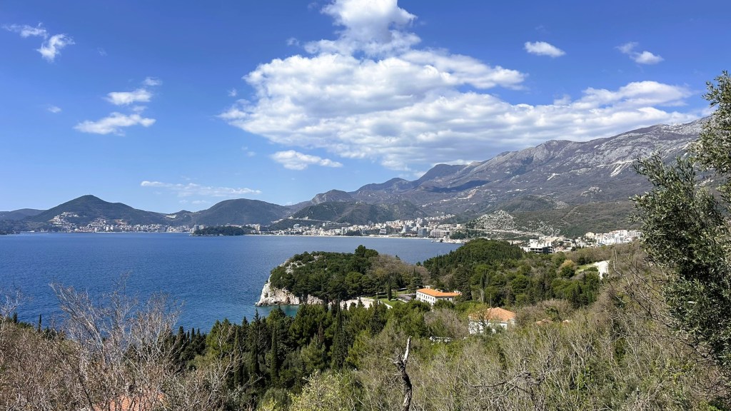 A scenic view of a coastal landscape featuring a calm blue sea, lush greenery, and mountains under a partly cloudy sky. Residential buildings are visible along the shoreline.