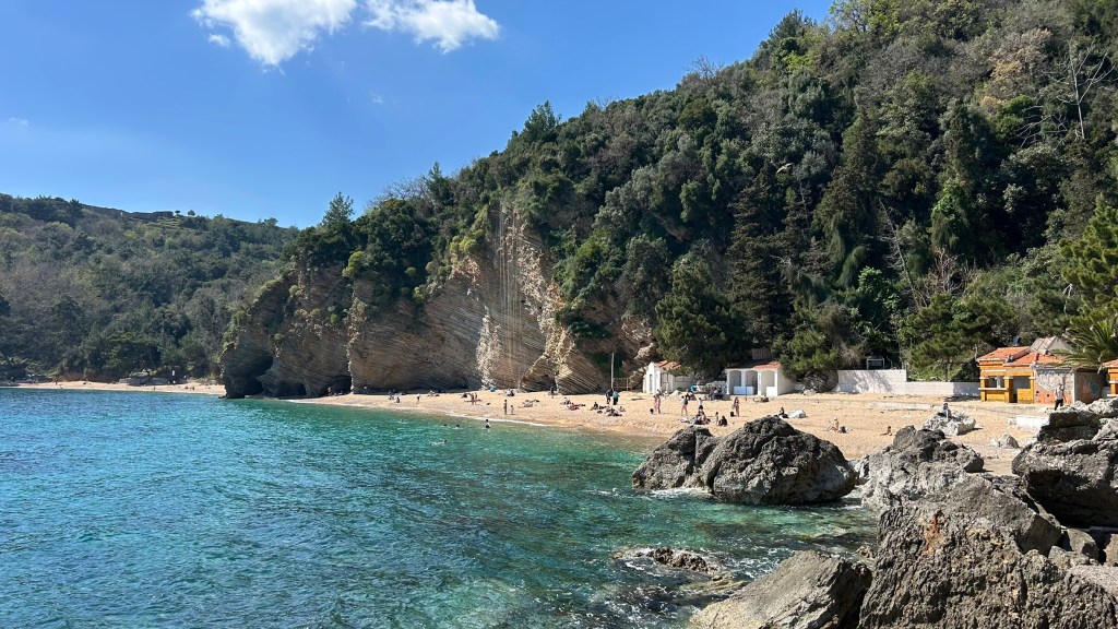 Scenic beach view with sandy shore, clear blue water, rocky foreground, and lush green hills in the background.