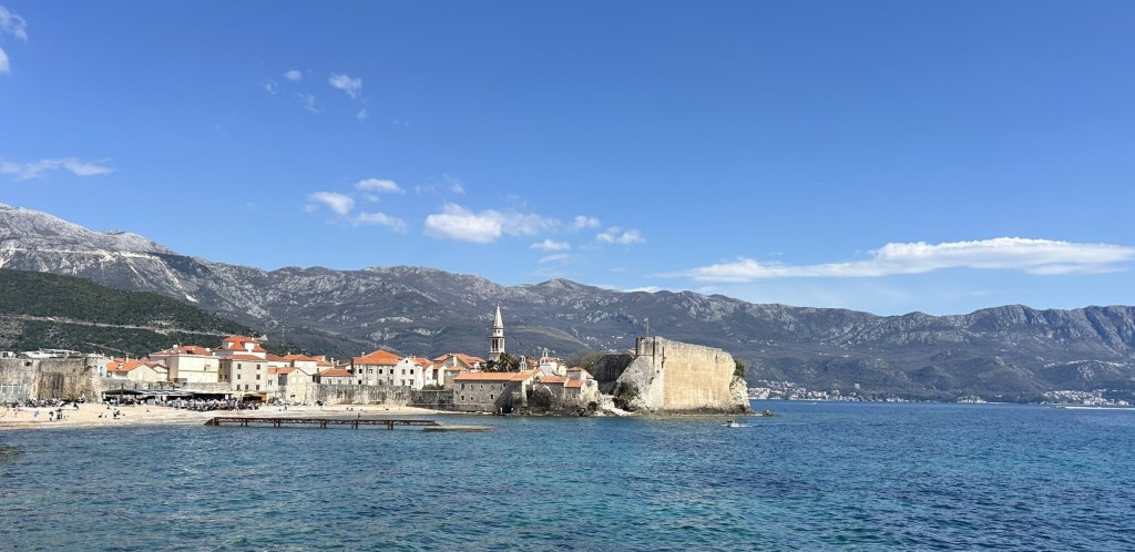 A scenic view of a coastal town with terracotta-roofed buildings, a stone fortress, and mountains in the background under a clear blue sky.