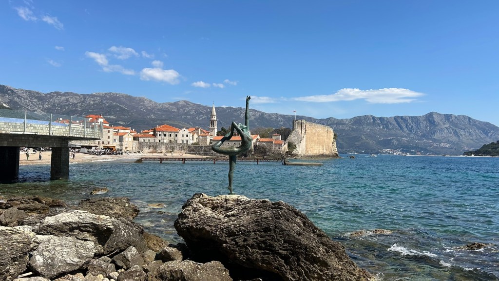 A coastal view featuring a sculpture of a dancer on a rock, with a historic town in the background, surrounded by mountains and clear blue water.