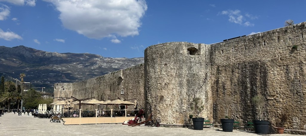 Historic stone city wall with a restaurant area in the foreground, set against a mountainous backdrop and blue sky.
