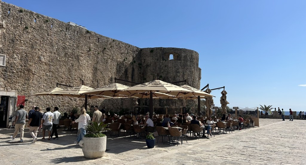 Outdoor café with seating under large umbrellas near a historic stone wall, with people walking by and a clear blue sky.