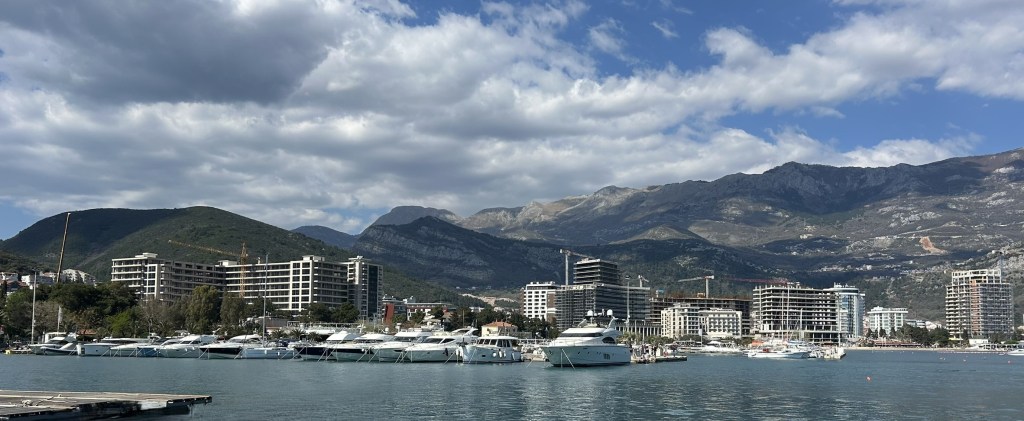 A scenic view of a marina with several yachts docked, surrounded by modern buildings and mountains under a partly cloudy sky.