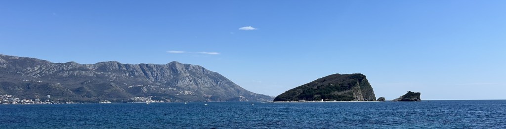 A panoramic view of a rocky coastline with two islands in the sea, backed by mountains and a clear blue sky.