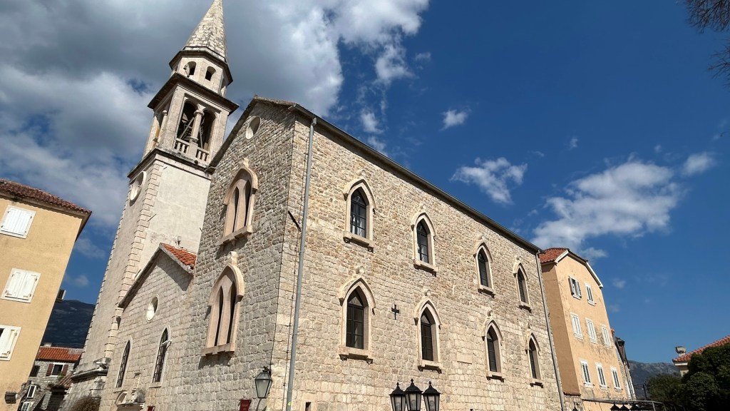 A stone church with a tall spire, featuring Gothic-style windows, set against a blue sky with scattered clouds.