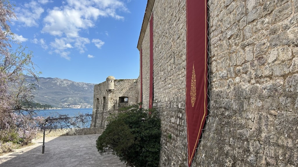 A view of a stone castle wall adorned with red banners, overlooking a lake with mountains in the background and a partly cloudy sky.